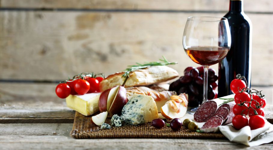 Picture of a chopping board with various cold cuts and fruit, a glass of wine and a bottle next to it.