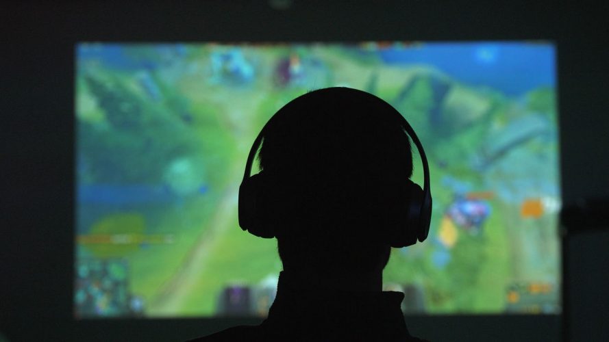 Silhouette di spalle di un uomo con delle cuffie che gioca con un videogame davanti a un grande schermo in una stanza scura