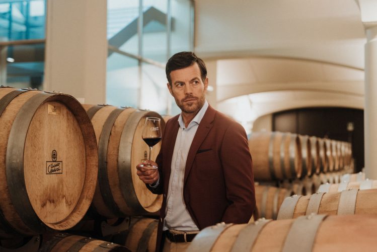 Un uomo ben vestito con una fila di botti alle sue spalle assaggia un vino rosso in una cantina