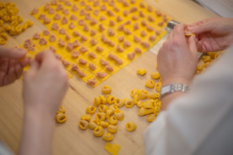 Primo piano delle mani di due persone che preparano dei tortellini, sullo sfondo il tavolo con file di tortellini da chiudere e alcuni tortellini già pronti